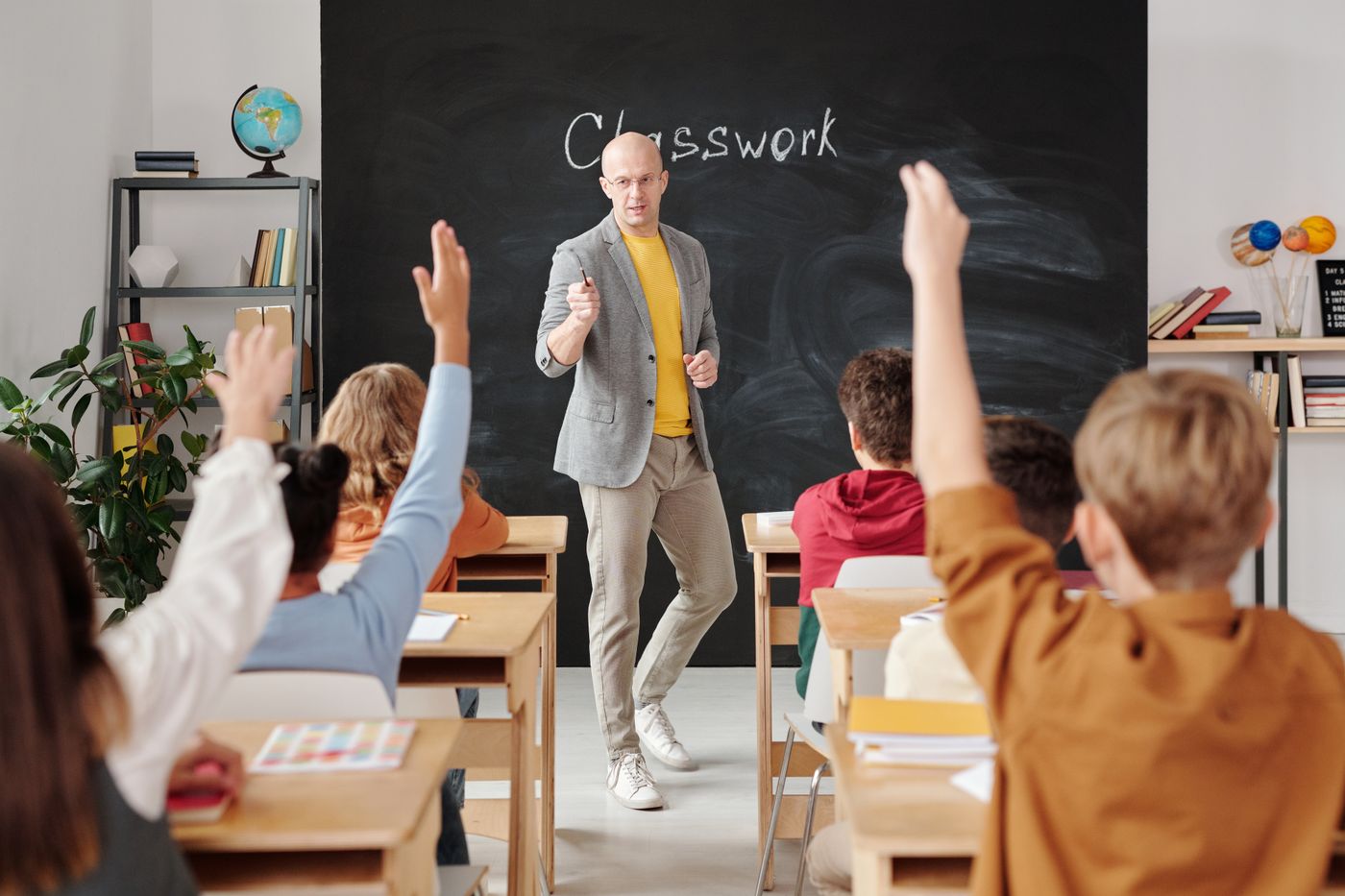 Teacher at a chalkboard labeled Classwork while students raise their hands