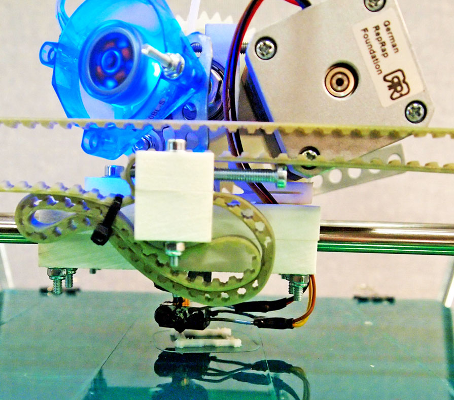 Close-up of a desktop 3D printer with belts, a blue fan, and a RepRap plate